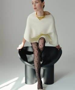 Model seated in a white poncho with gold jewelry, wearing black heart-pattern tights on a modern gray chair in a studio.