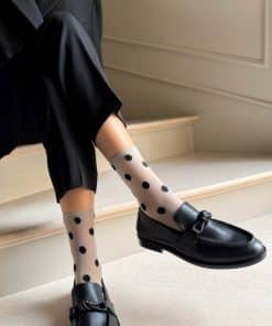 Person seated on light stairs wearing black loafers and sheer white socks with large black polka dots, paired with black pants.