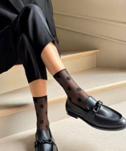 Person wearing sheer black socks with large polka dots and black loafers, seated on light stairs in a black suit.