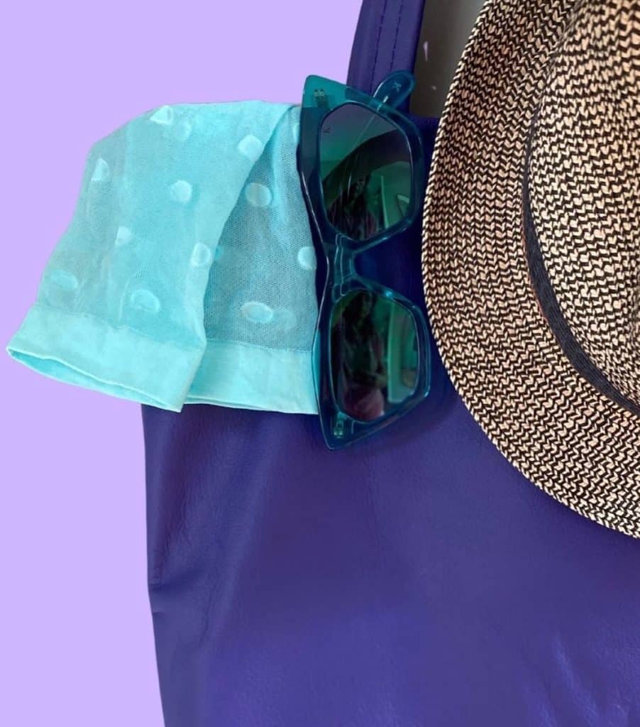 Purple bag with a turquoise scarf, teal sunglasses hanging on the edge, and a woven straw hat resting on the side, set against a lavender background.