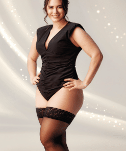 Confident woman in black ruched bodysuit and thigh-high stockings posing with hands on hips, smiling against a light, sparkling background.